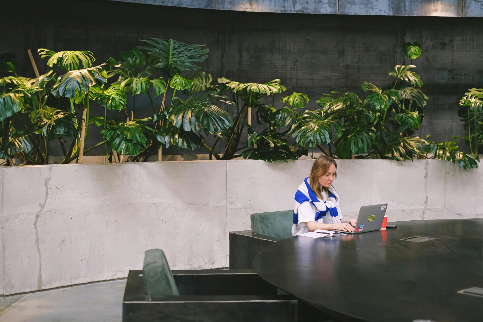 Woman working on a laptop in a modern indoor setting with lush greenery and sleek furniture.
