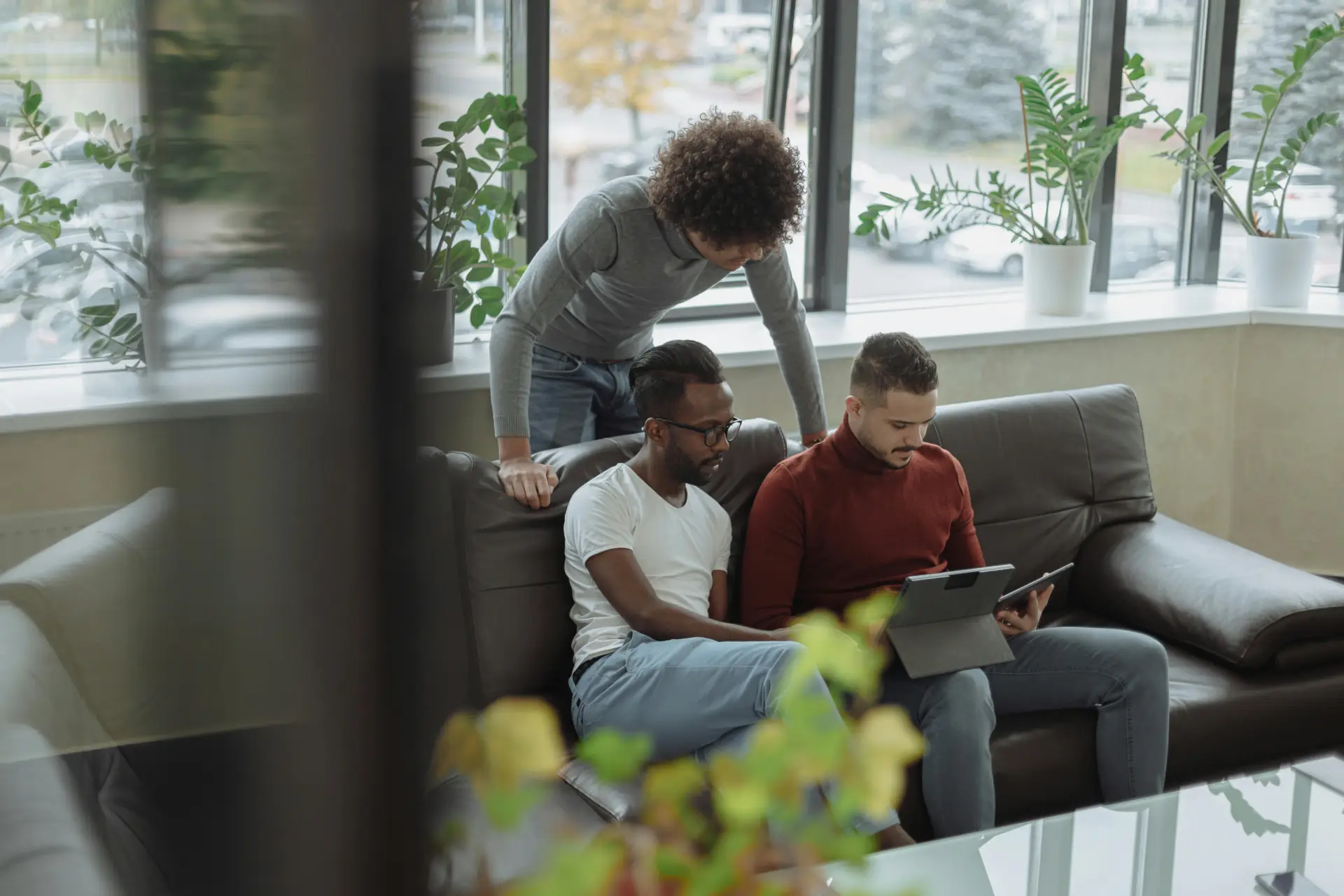 Three men discuss projects using a tablet in a bright office lounge, promoting teamwork.
