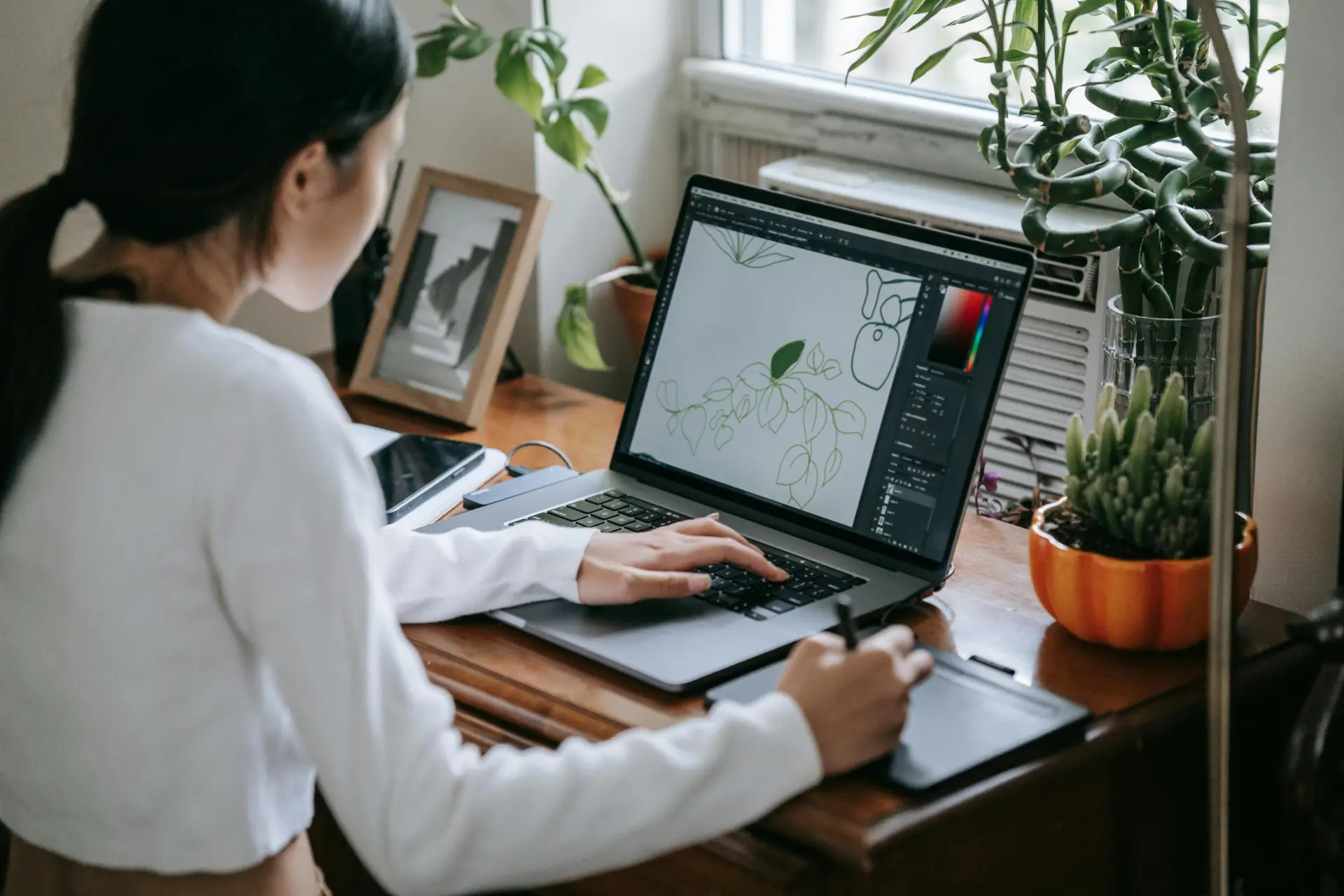 Woman using a stylus to draw on a laptop at a desk, surrounded by plants indoors.
