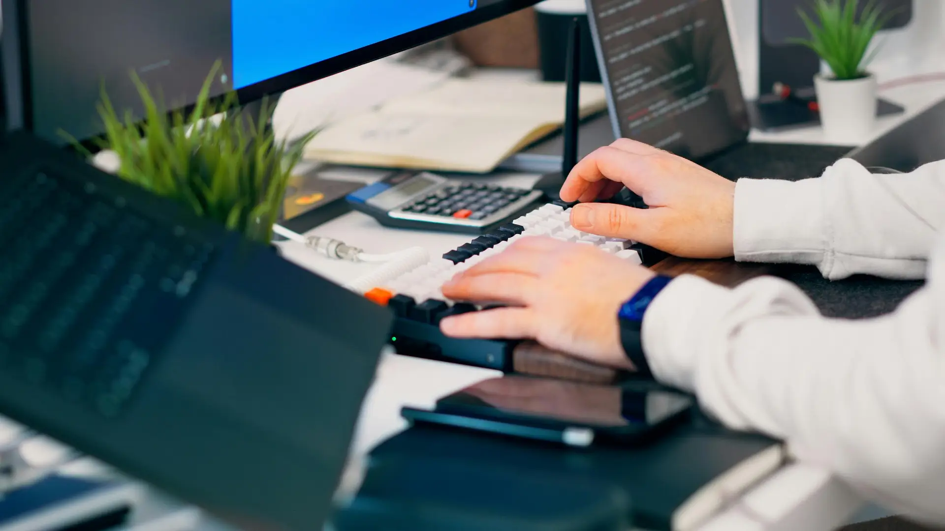 A close-up of a person typing on a keyboard in a modern tech workspace with gadgets and a monitor.
