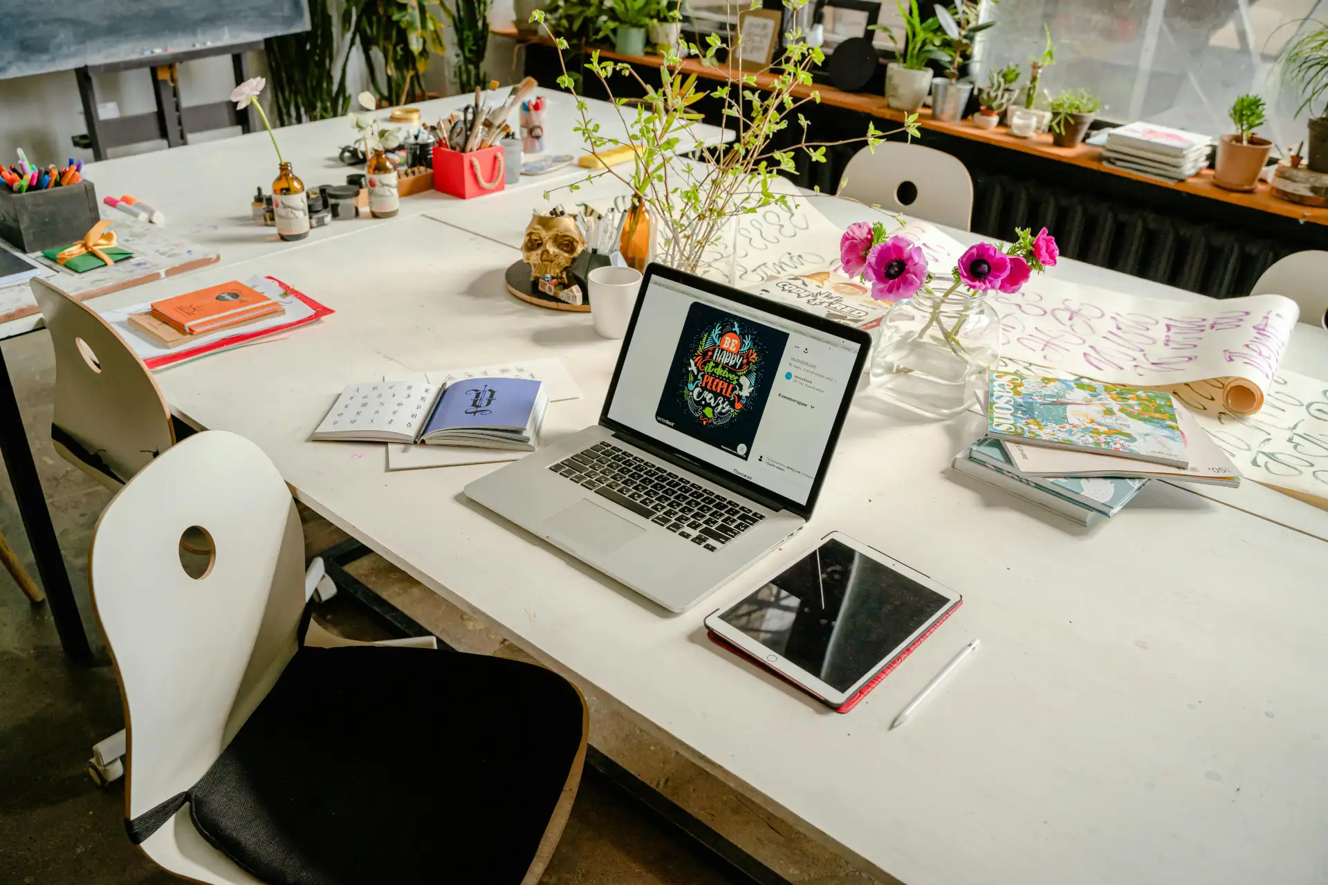 Artistic workspace featuring a laptop, tablet, flowers, and art supplies on a table.
