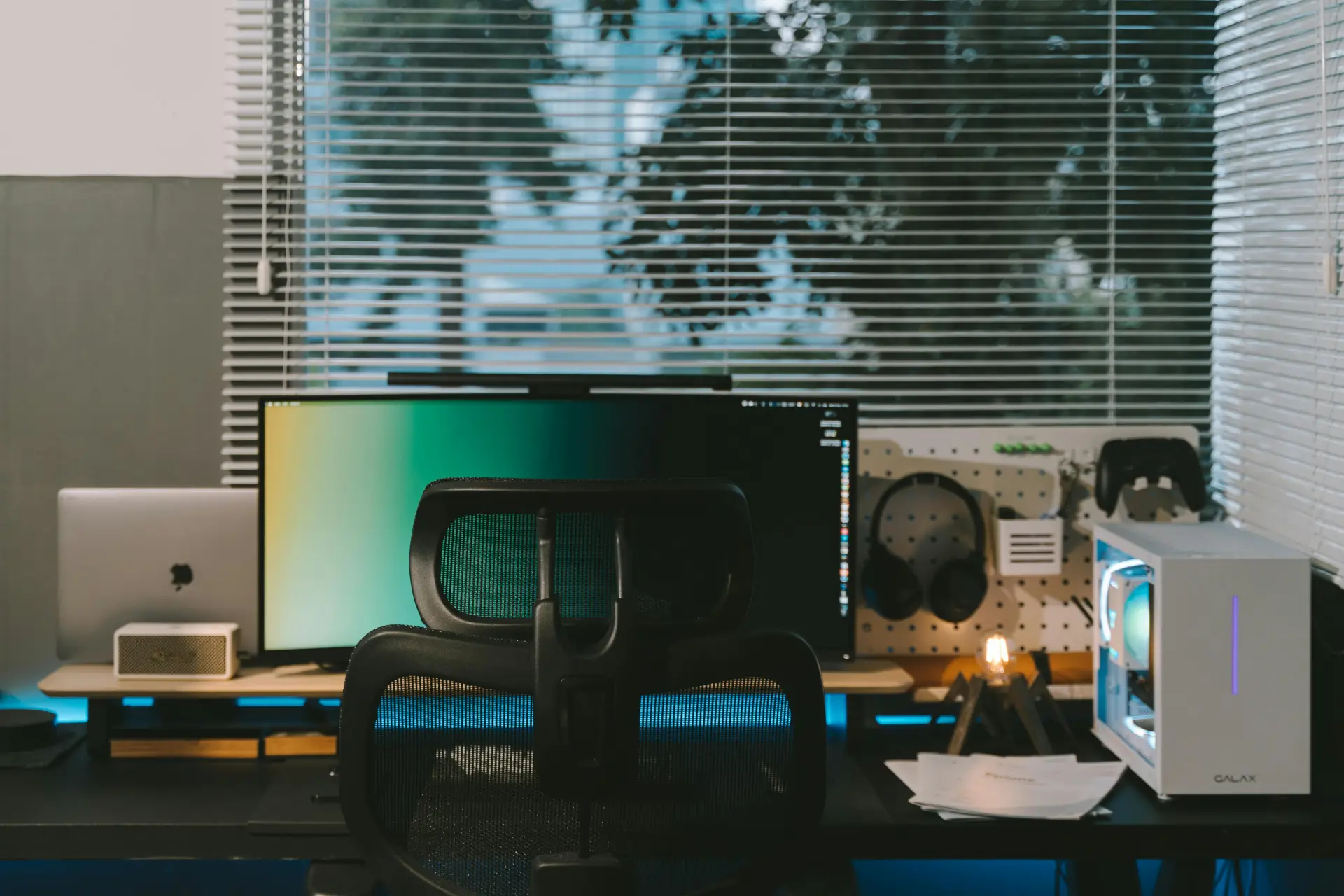 A stylish office desk setup featuring a computer, gadgets, and large windows with blinds.
