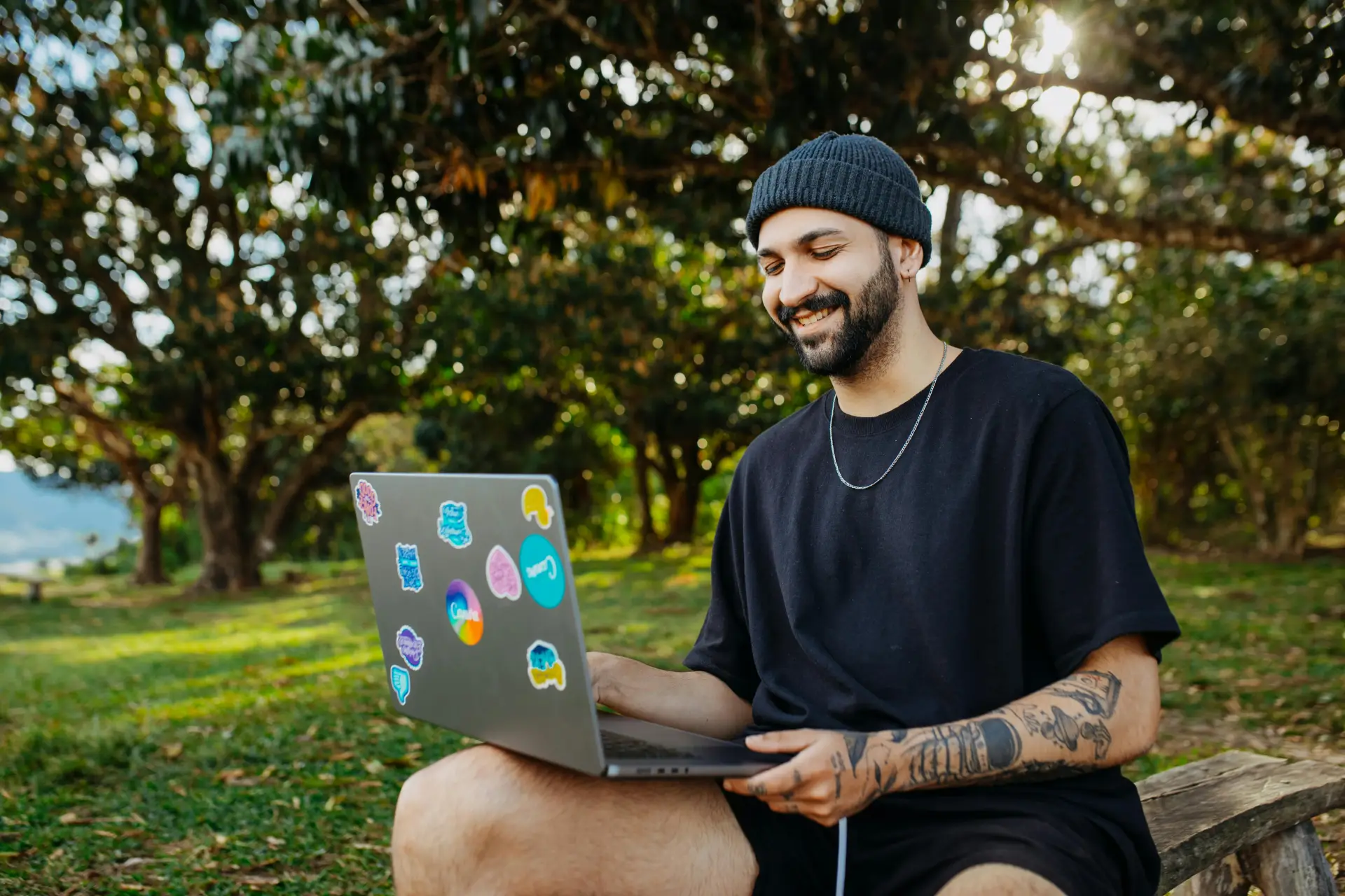 A smiling man with tattoos working on a laptop outdoors in a sunny park setting.
