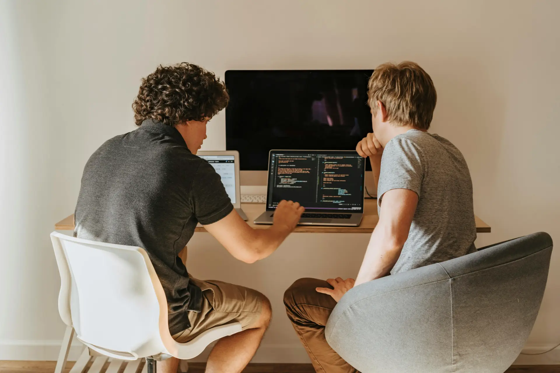 Two male developers working on laptops indoors, discussing code.
