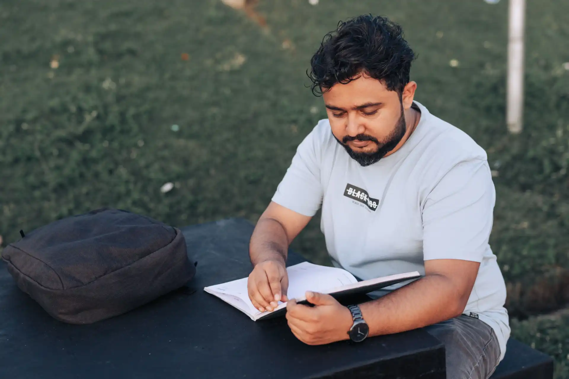 A young man in casual attire studying outdoors with a notebook and backpack.