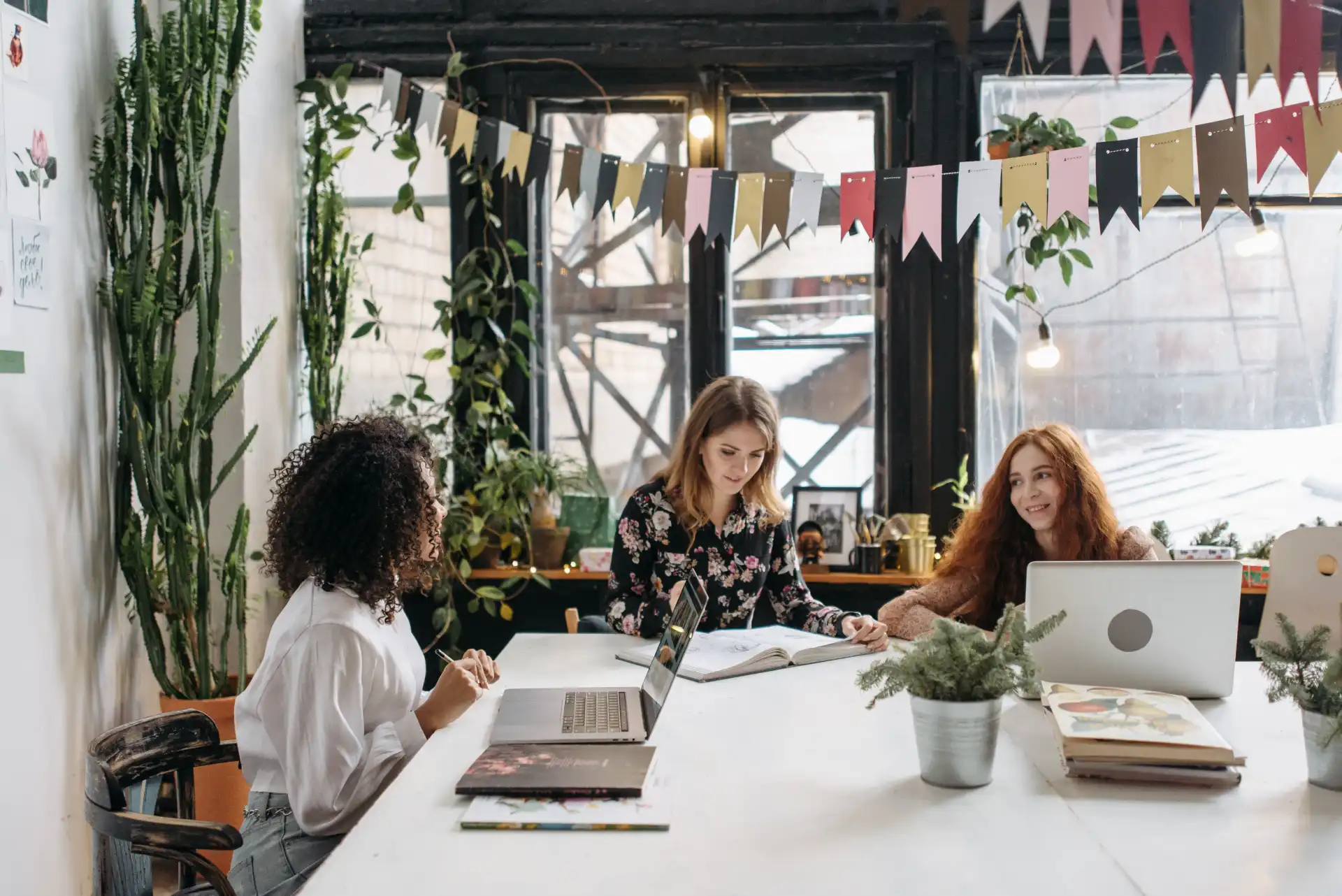 Three women working collaboratively in a stylish, plant-filled workspace featuring laptops, bunting, and natural light.
