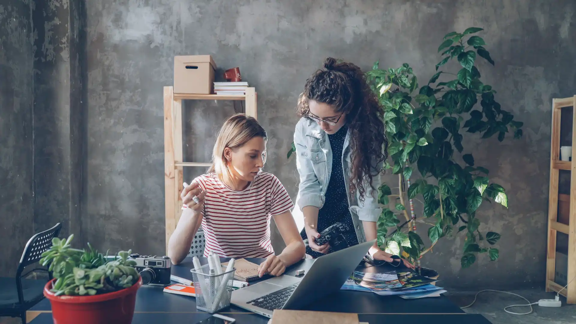 Two women discussing photography in a stylish loft workspace with plants.

