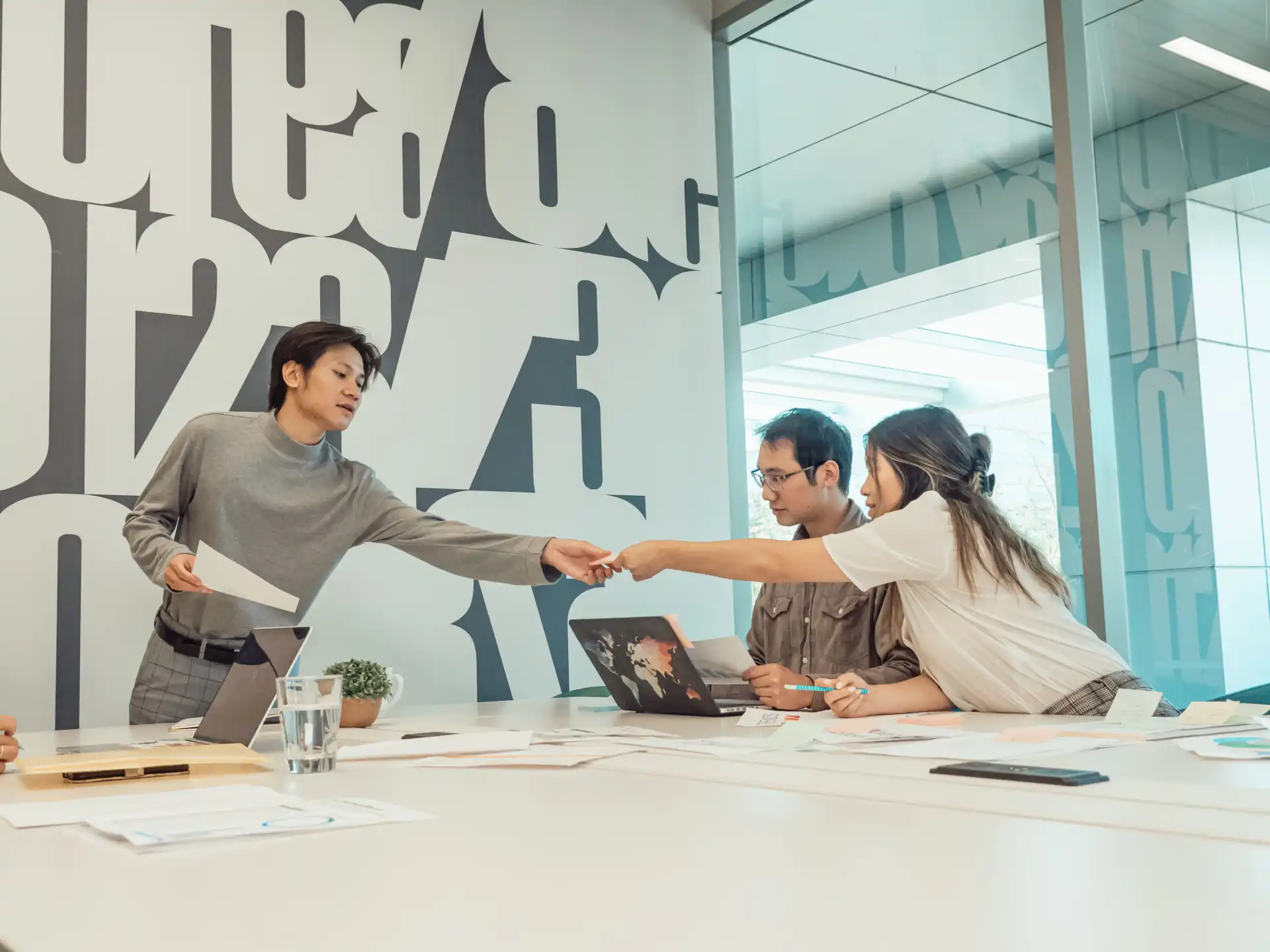 Three coworkers engaged in a teamwork session, exchanging documents over a conference table.
